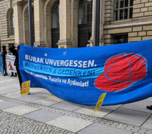 Kundgebung vor dem Abgeordnetenhaus Berlin. Zu sehen ist ein blaues Banner mit der Aufschrift: "Burak unvergessen. Aufklären & Gedenken! Burak´ı unutma. Hatırla ve Aydınlat!" Auf dem Banner ist eine stilisierte rote Kappe dargestellt.